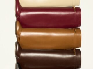 Cinco botas de cano longo em couro empilhadas nas cores creme, vinho, caramelo, marrom e preto, com detalhe metálico dourado no cano