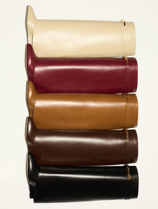 Cinco botas de cano longo em couro empilhadas nas cores creme, vinho, caramelo, marrom e preto, com detalhe metálico dourado no cano