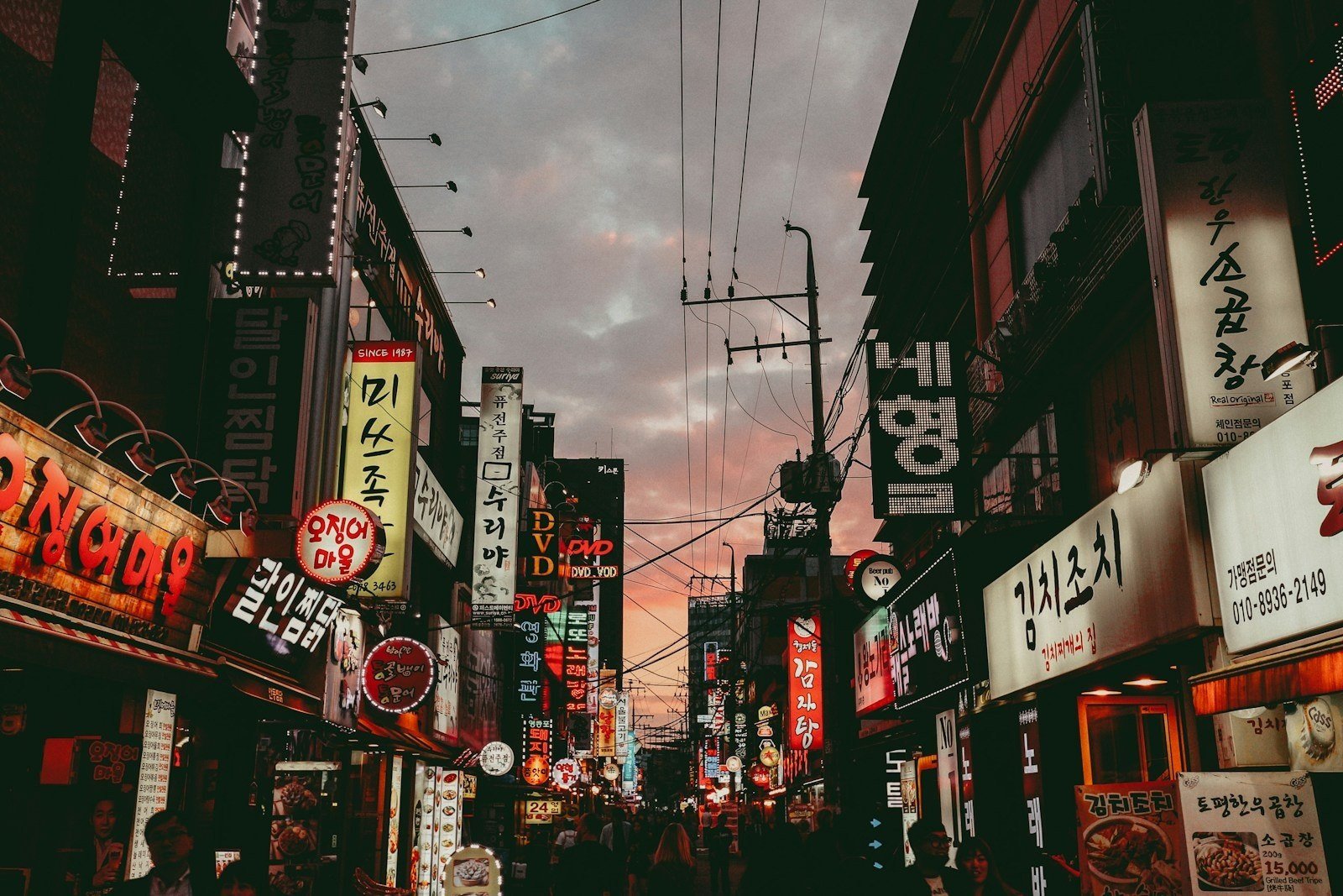 Rua comercial de Seul ao entardecer, com letreiros luminosos em coreano dos dois lados da via