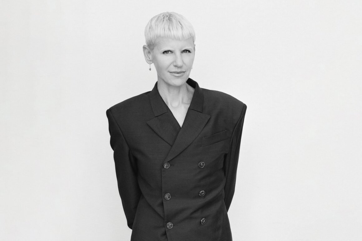 Retrato em preto e branco de Erin Magee, de cabelo curto platinado e blazer escuro de ombros largos, contra fundo claro