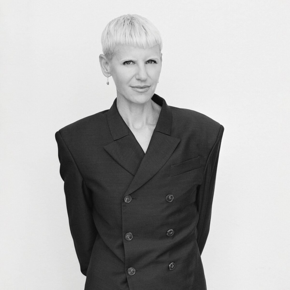 Retrato em preto e branco de Erin Magee, de cabelo curto platinado e blazer escuro de ombros largos, contra fundo claro