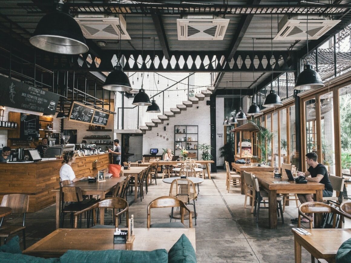 Salão amplo de restaurante com mesas e cadeiras de madeira, luminárias industriais pretas e grandes janelas