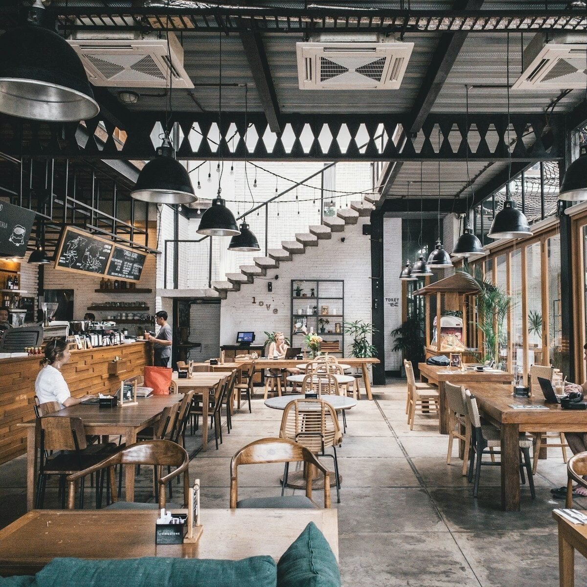 Salão amplo de restaurante com mesas e cadeiras de madeira, luminárias industriais pretas e grandes janelas