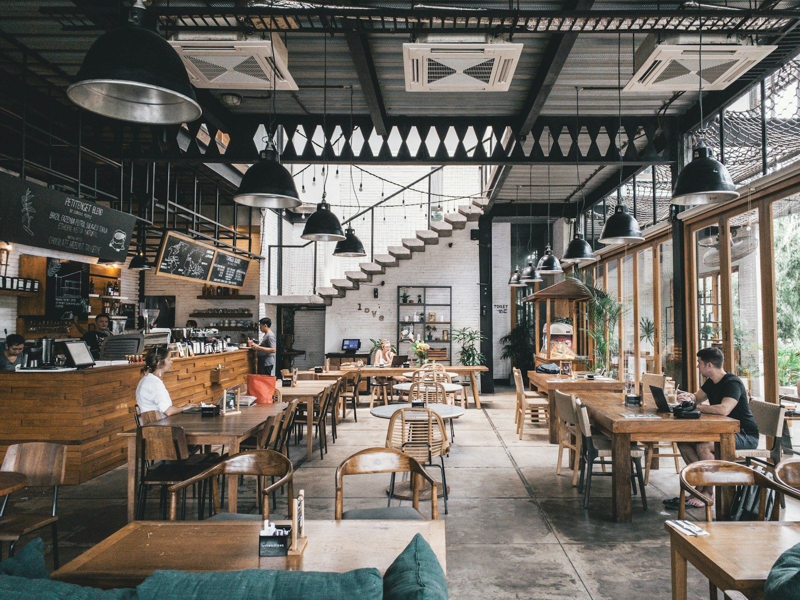 Salão amplo de restaurante com mesas e cadeiras de madeira, luminárias industriais pretas e grandes janelas