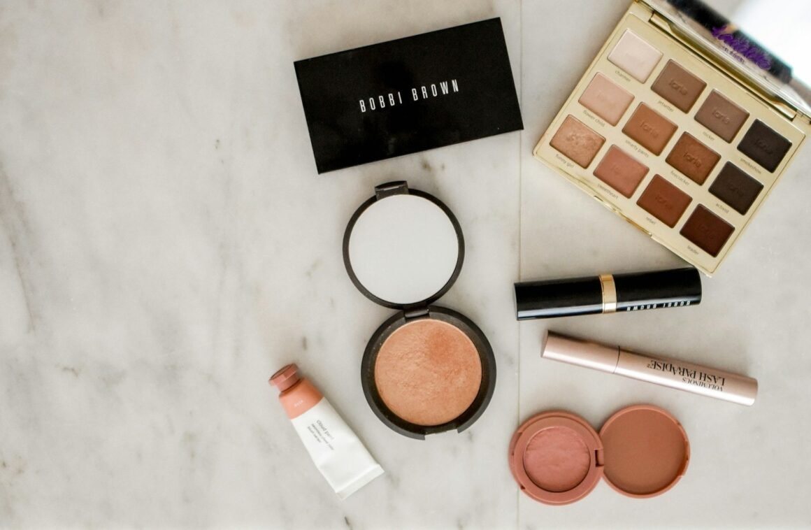 Produtos de maquiagem sobre superfície de mármore: paletas de sombra, iluminador, batom, máscara de cílios e blush
