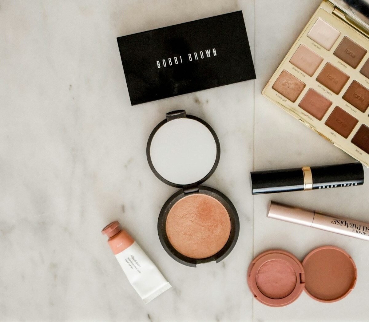 Produtos de maquiagem sobre superfície de mármore: paletas de sombra, iluminador, batom, máscara de cílios e blush