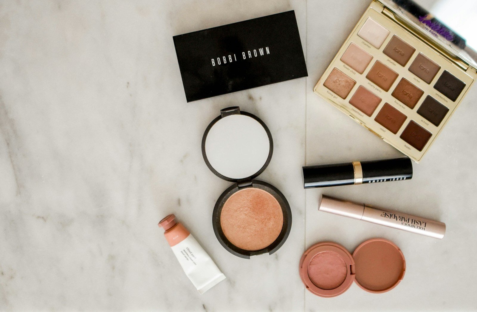 Produtos de maquiagem sobre superfície de mármore: paletas de sombra, iluminador, batom, máscara de cílios e blush