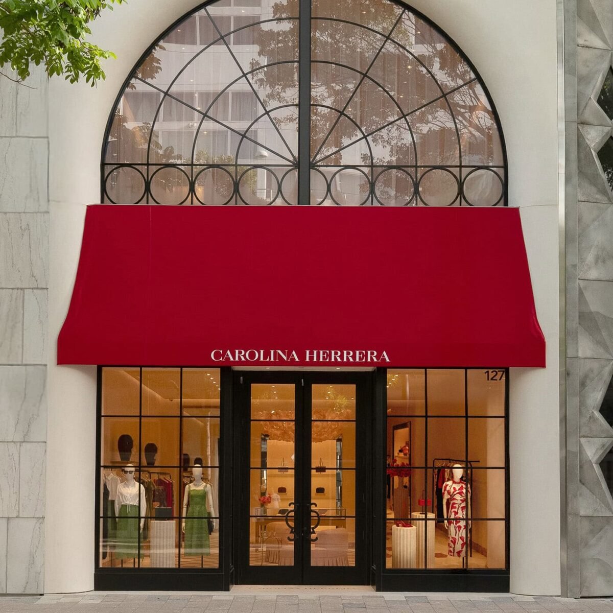 Entrada da loja Carolina Herrera com toldo vermelho sob arco de vidro e vitrine com manequins de vestidos verdes e vermelhos