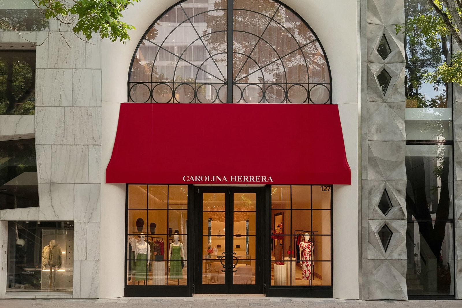 Entrada da loja Carolina Herrera com toldo vermelho sob arco de vidro e vitrine com manequins de vestidos verdes e vermelhos