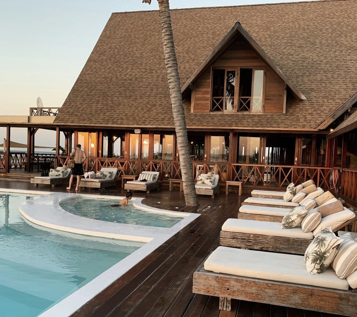 Piscina e espreguiçadeiras em deck de madeira diante de hotel de praia ao entardecer
