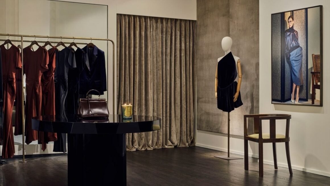 Interior da loja com arara de vestidos, bolsa sobre mesa central preta, manequim com vestido de veludo e quadro na parede