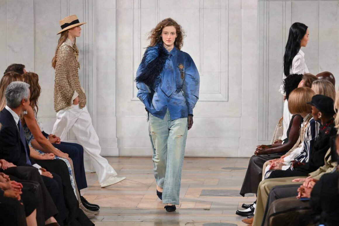 Modelo desfila com jaqueta azul de cetim e plumas sobre jeans claro em passarela de salão clássico, com plateia nas laterais