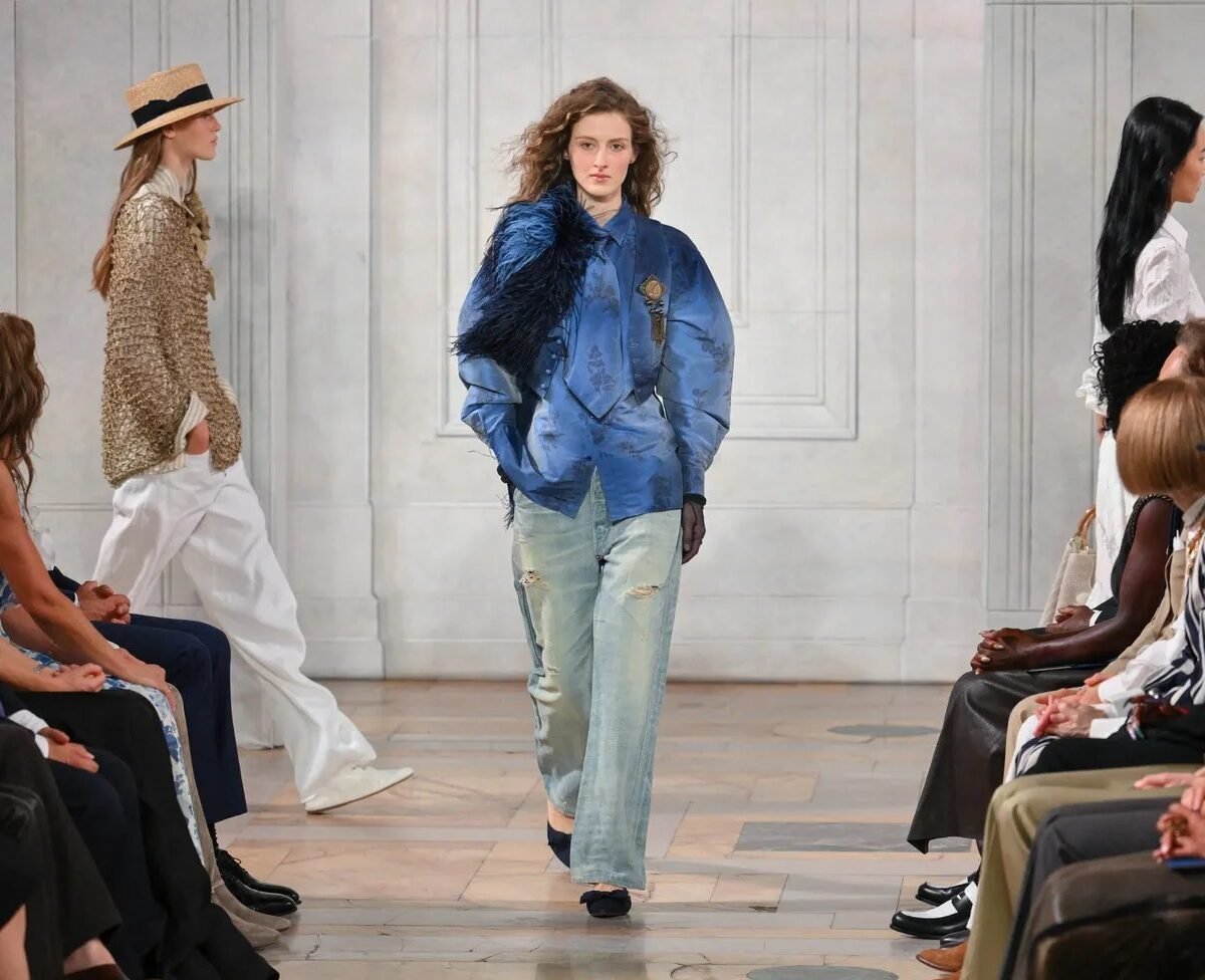 Modelo desfila com jaqueta azul de cetim e plumas sobre jeans claro em passarela de salão clássico, com plateia nas laterais