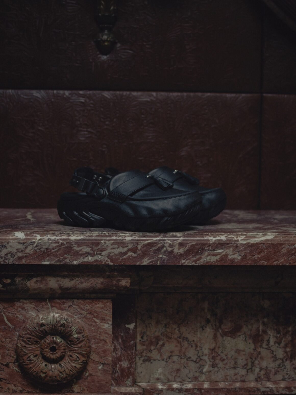 Clog de couro preto com faixa frontal, tassel e fivela traseira, apoiado sobre lareira de mármore