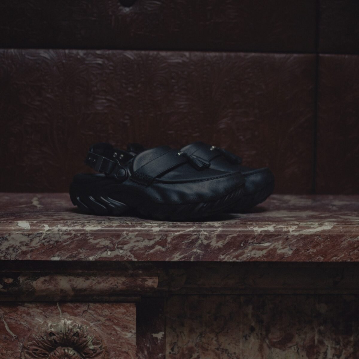 Clog de couro preto com faixa frontal, tassel e fivela traseira, apoiado sobre lareira de mármore