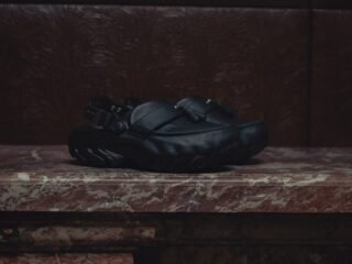 Clog de couro preto com faixa frontal, tassel e fivela traseira, apoiado sobre lareira de mármore