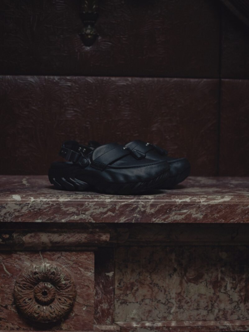 Clog de couro preto com faixa frontal, tassel e fivela traseira, apoiado sobre lareira de mármore