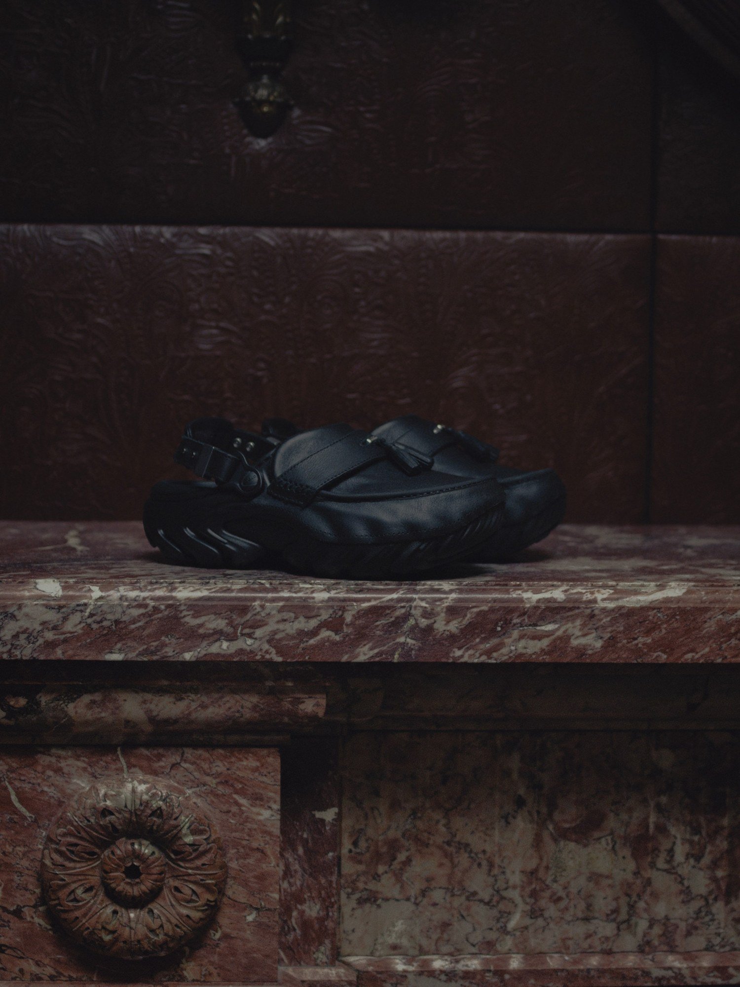 Clog de couro preto com faixa frontal, tassel e fivela traseira, apoiado sobre lareira de mármore