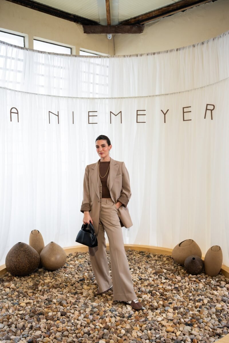 Convidada de terno e calça camel, blusa marrom e bolsa preta, diante do painel de tecido branco com o nome A NIEMEYER, sobre um círculo de pedras