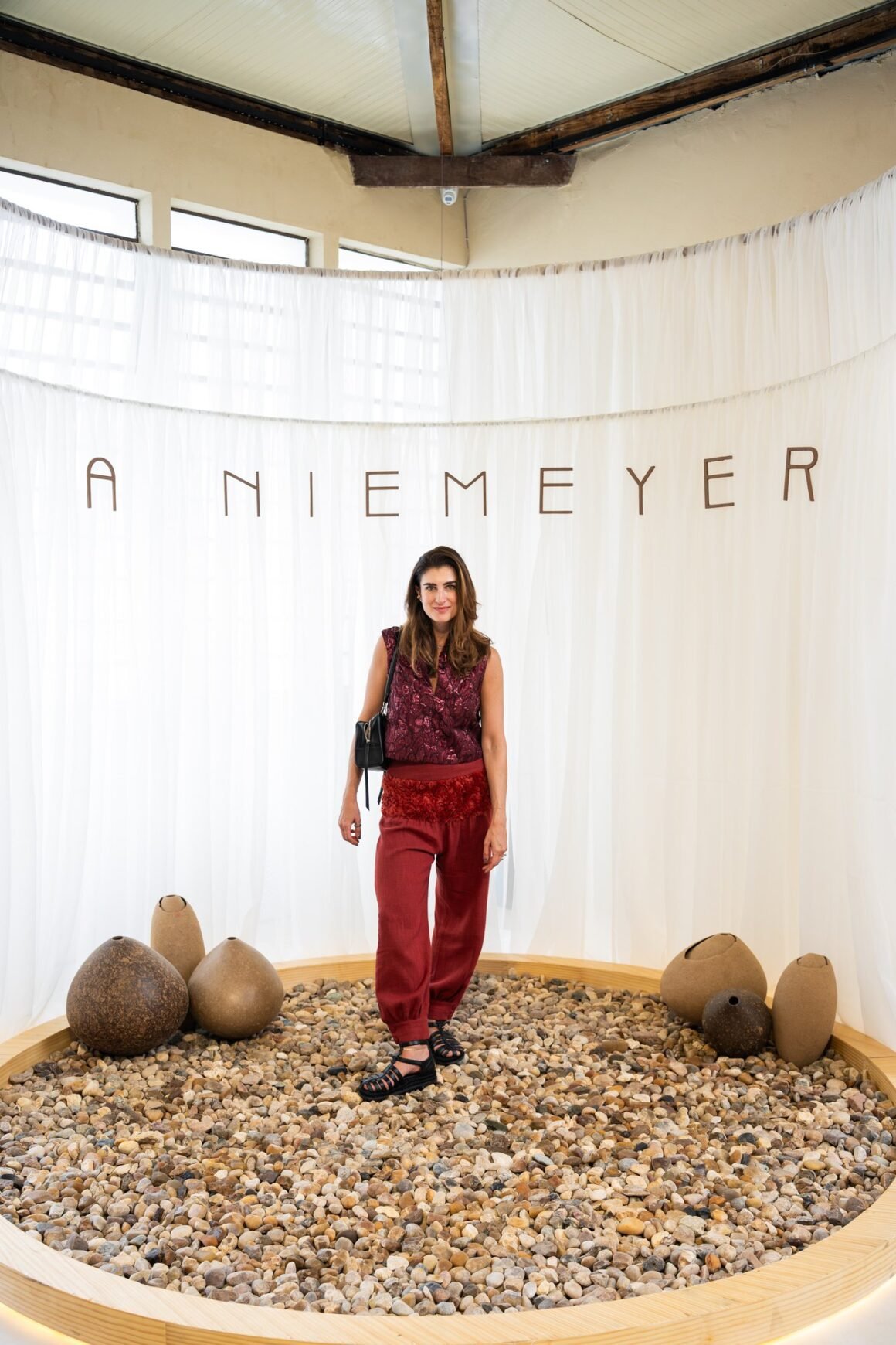 Convidada de blusa e calça vermelhas em pé sobre o círculo de pedras diante do painel de tecido branco com o nome A NIEMEYER