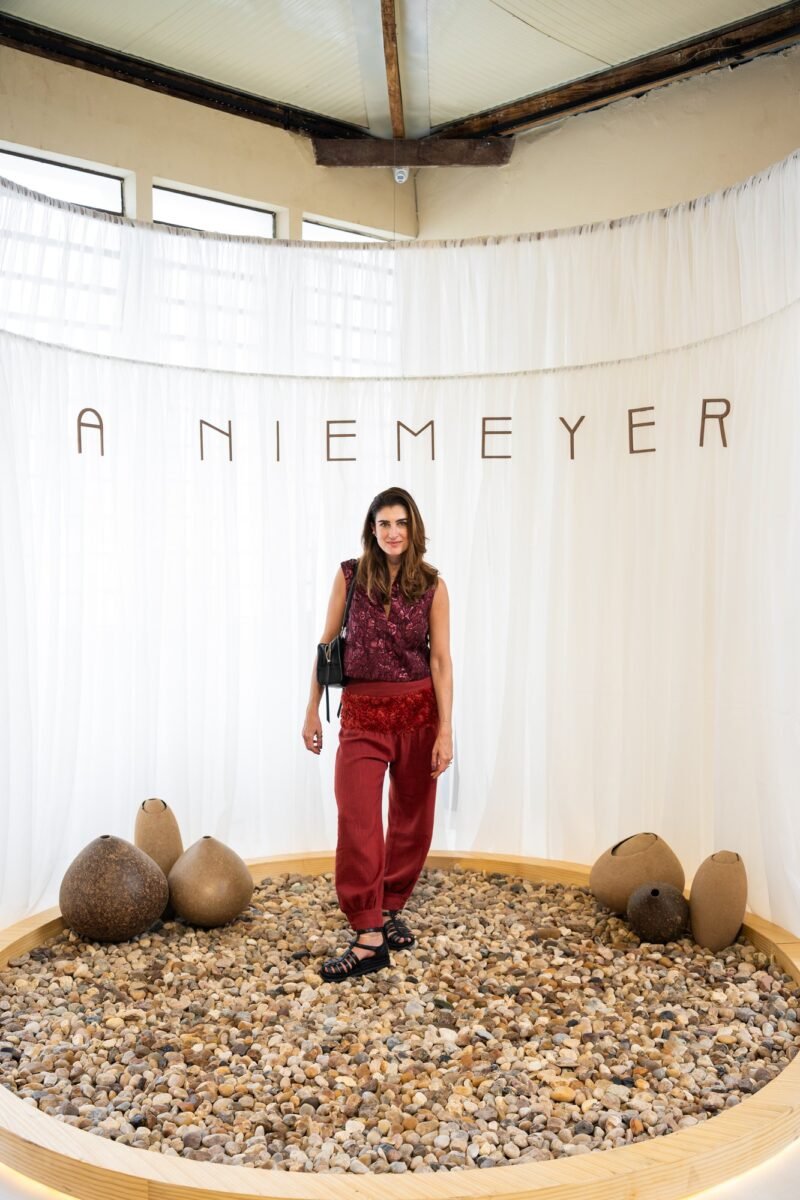 Convidada de blusa e calça vermelhas em pé sobre o círculo de pedras diante do painel de tecido branco com o nome A NIEMEYER