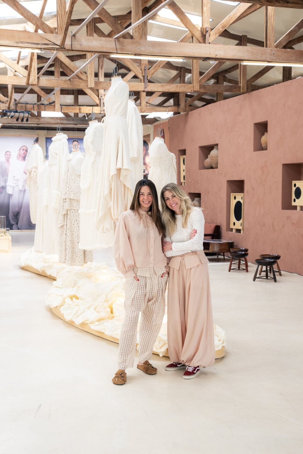 Duas mulheres abraçadas no galpão da exposição, diante de araras com peças em off-white e de uma parede de terracota com nichos