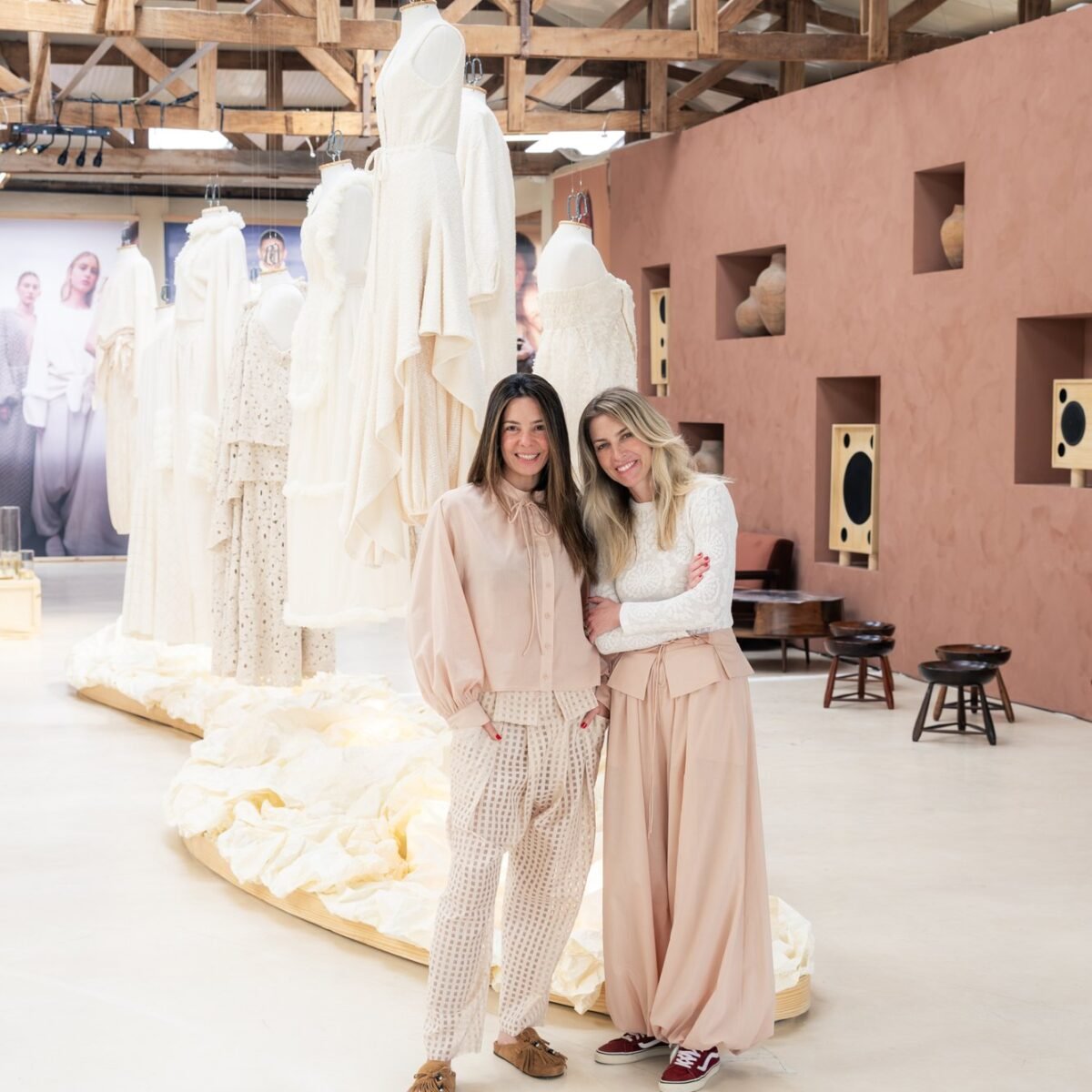 Duas mulheres abraçadas no galpão da exposição, diante de araras com peças em off-white e de uma parede de terracota com nichos