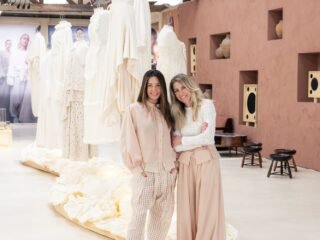 Duas mulheres abraçadas no galpão da exposição, diante de araras com peças em off-white e de uma parede de terracota com nichos