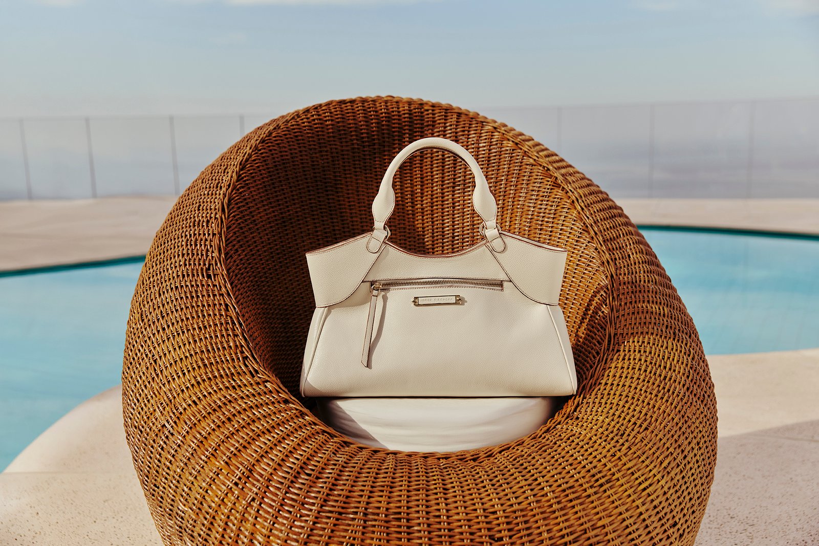 Bolsa branca da coleção Rico por Natureza, da Jorge Bischoff, sobre poltrona de fibra natural à beira da piscina