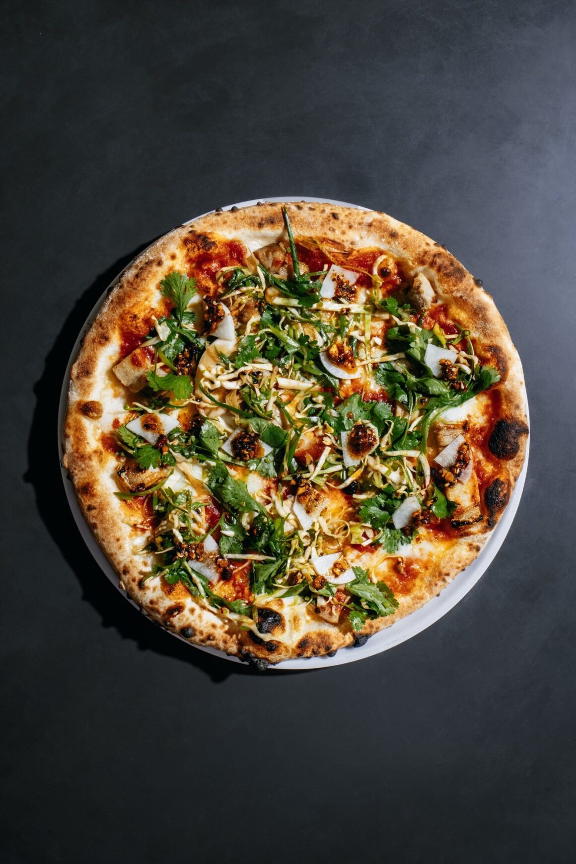 Pizza redonda de borda tostada coberta com folhas verdes, salada de repolho, picles e pimenta, vista de cima sobre fundo escuro