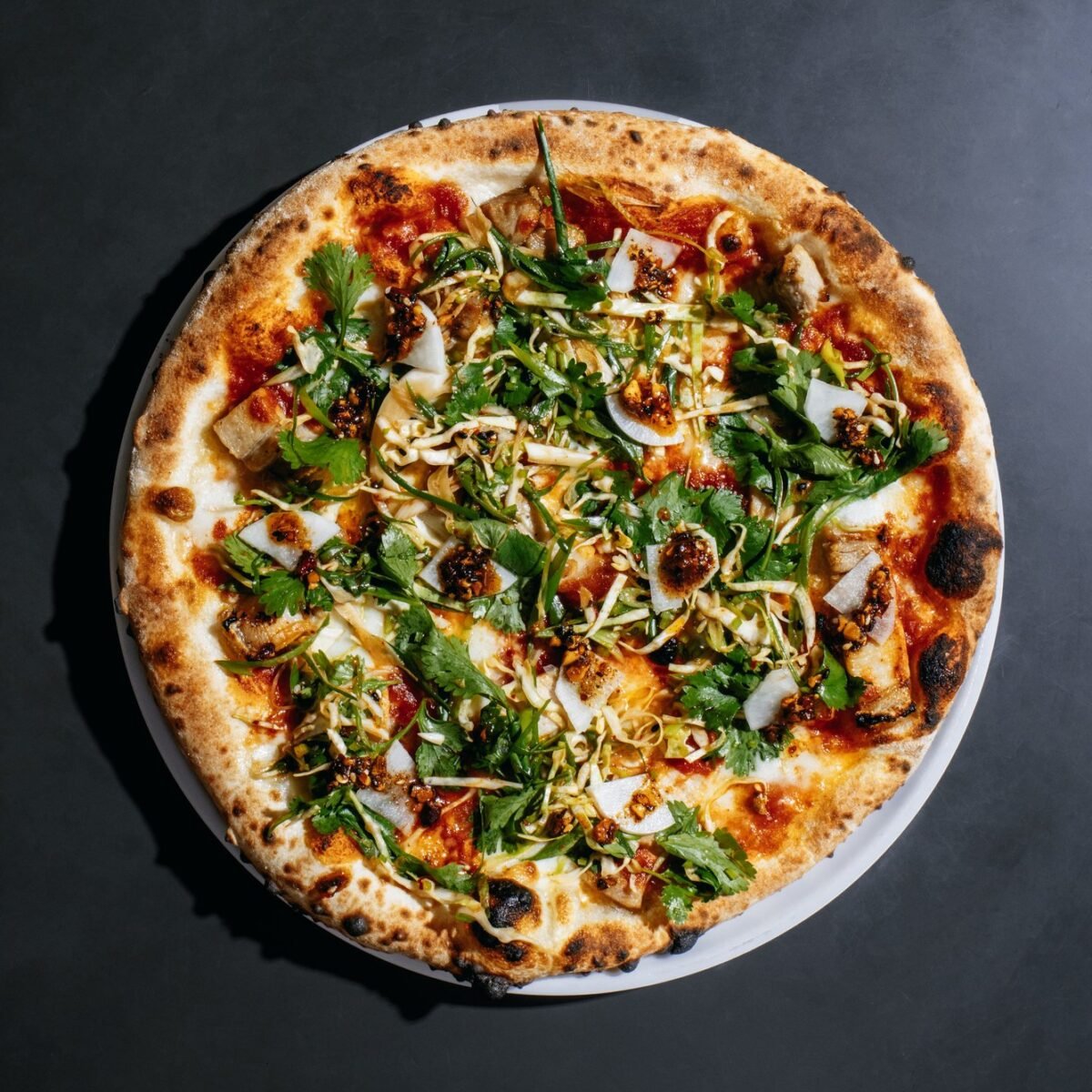 Pizza redonda de borda tostada coberta com folhas verdes, salada de repolho, picles e pimenta, vista de cima sobre fundo escuro