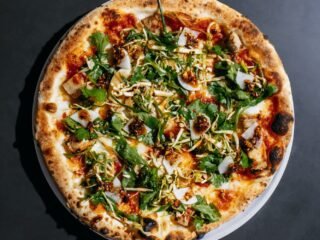 Pizza redonda de borda tostada coberta com folhas verdes, salada de repolho, picles e pimenta, vista de cima sobre fundo escuro