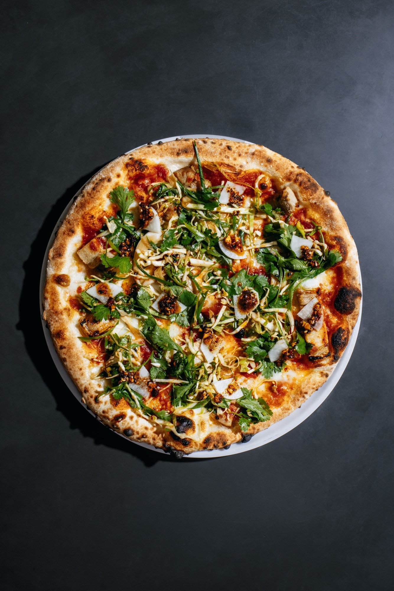 Pizza redonda de borda tostada coberta com folhas verdes, salada de repolho, picles e pimenta, vista de cima sobre fundo escuro