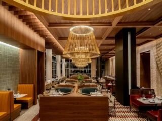Salão de restaurante com forro de madeira ripada, grandes luminárias de palha, bancos estofados em laranja e vermelho e tapete geométrico