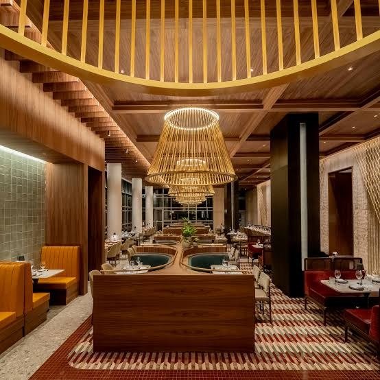 Salão de restaurante com forro de madeira ripada, grandes luminárias de palha, bancos estofados em laranja e vermelho e tapete geométrico