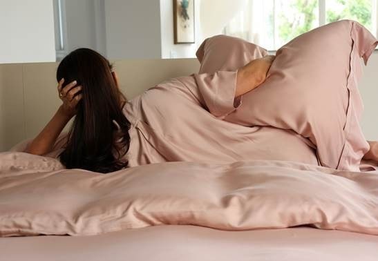 Cama com jogo de roupa de cama em rosa acetinado, com edredom e fronhas, e uma mulher deitada de lado apoiando o rosto na mão