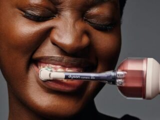 Mulher escova os dentes com a escova elétrica da Dyson, cuja haste traz uma câmera acoplada, em fundo cinza