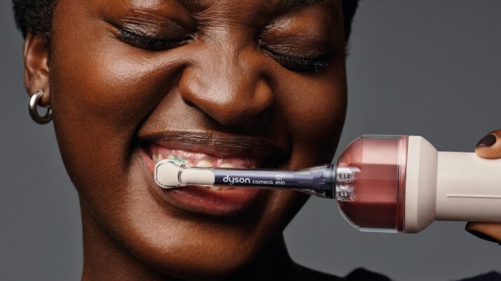 Mulher escova os dentes com a escova elétrica da Dyson, cuja haste traz uma câmera acoplada, em fundo cinza