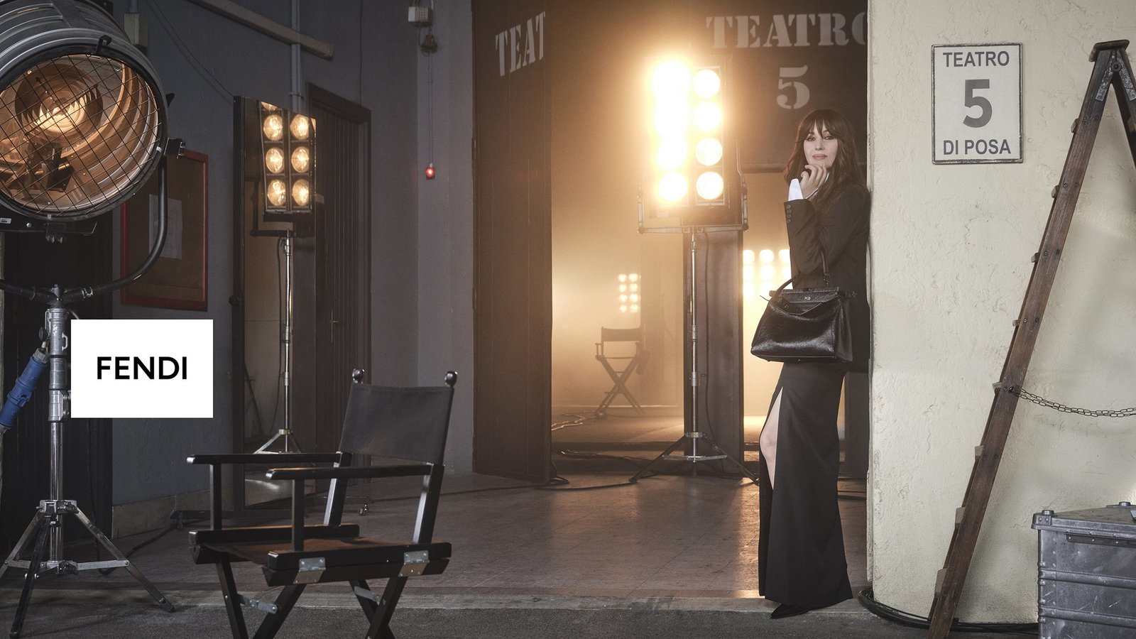 Monica Bellucci de terno preto e saia longa com fenda, apoiada em uma parede ao lado da placa Teatro 5 di Posa, com uma bolsa Peekaboo preta no braço, em set de filmagem com refletores acesos e cadeira de diretor em primeiro plano