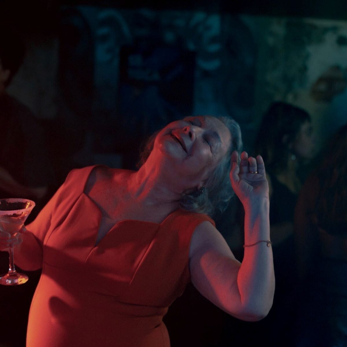 Mulher de vestido vermelho dança de olhos fechados segurando uma taça, sob luz vermelha e azul em ambiente noturno