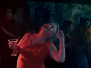 Mulher de vestido vermelho dança de olhos fechados segurando uma taça, sob luz vermelha e azul em ambiente noturno