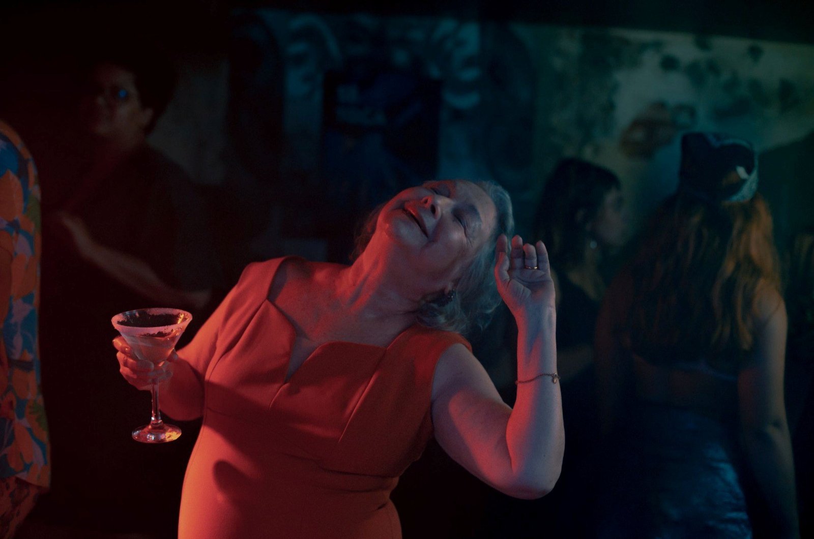 Mulher de vestido vermelho dança de olhos fechados segurando uma taça, sob luz vermelha e azul em ambiente noturno