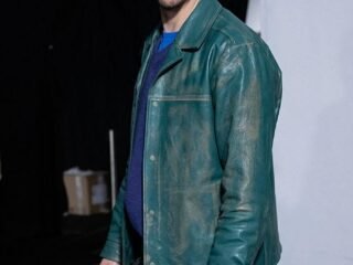 Homem de boné escuro e jaqueta de couro verde envelhecida sobre suéter azul posa nos bastidores de um desfile