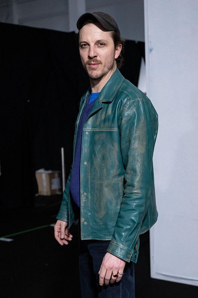 Homem de boné escuro e jaqueta de couro verde envelhecida sobre suéter azul posa nos bastidores de um desfile