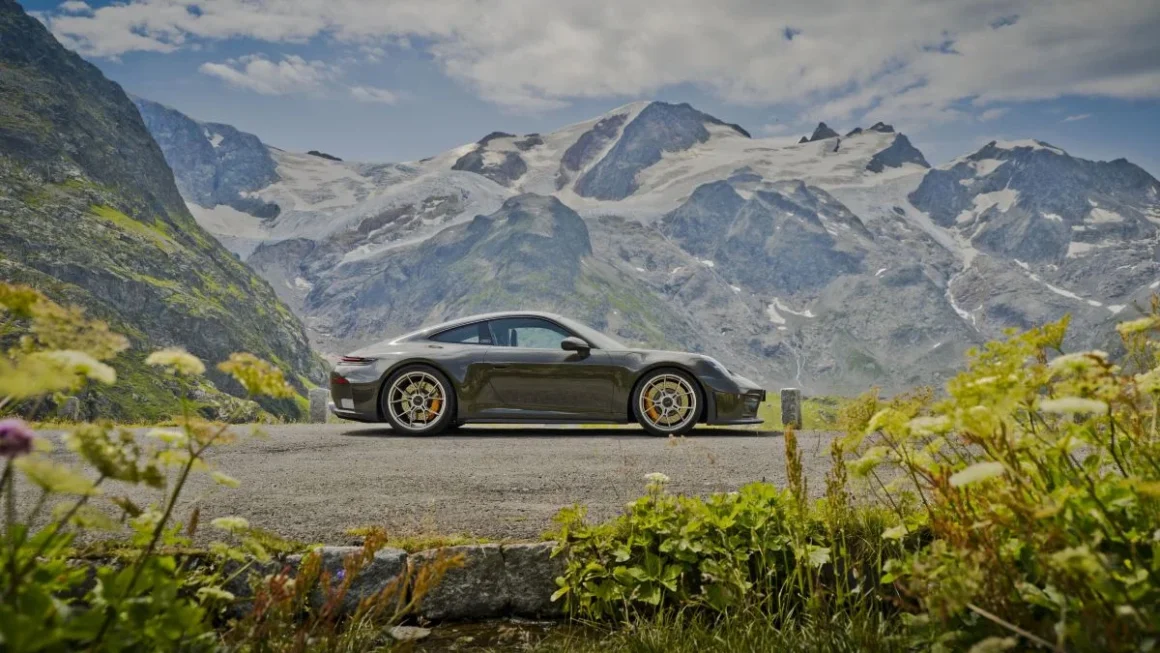 Porsche 911 GT3 cinza-esverdeado de perfil, parado em estrada de montanha, com picos nevados dos Alpes ao fundo e vegetação em primeiro plano