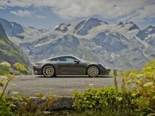Porsche 911 GT3 cinza-esverdeado de perfil, parado em estrada de montanha, com picos nevados dos Alpes ao fundo e vegetação em primeiro plano