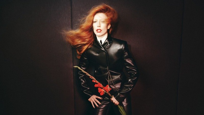Modelo ruiva de terno de couro preto segurando uma flor vermelha diante de uma parede escura.