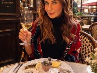 Mulher sentada em mesa de bistrô parisiense com taça de vinho branco e prato de ostras.