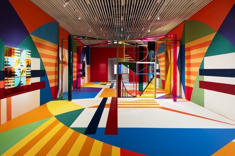 Sala de exposição com paredes e piso pintados em formas geométricas coloridas e estruturas de vidro.