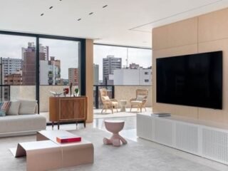 Sala de estar com painel de madeira clara, sofá cinza e banco Toy em rosa ao lado de mesa de centro baixa em tom blush, com varanda e vista da cidade ao fundo